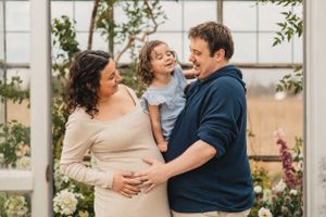 A small family embraces and laughs together during a romantic greenhouse photoshoot maternity photoshoot.