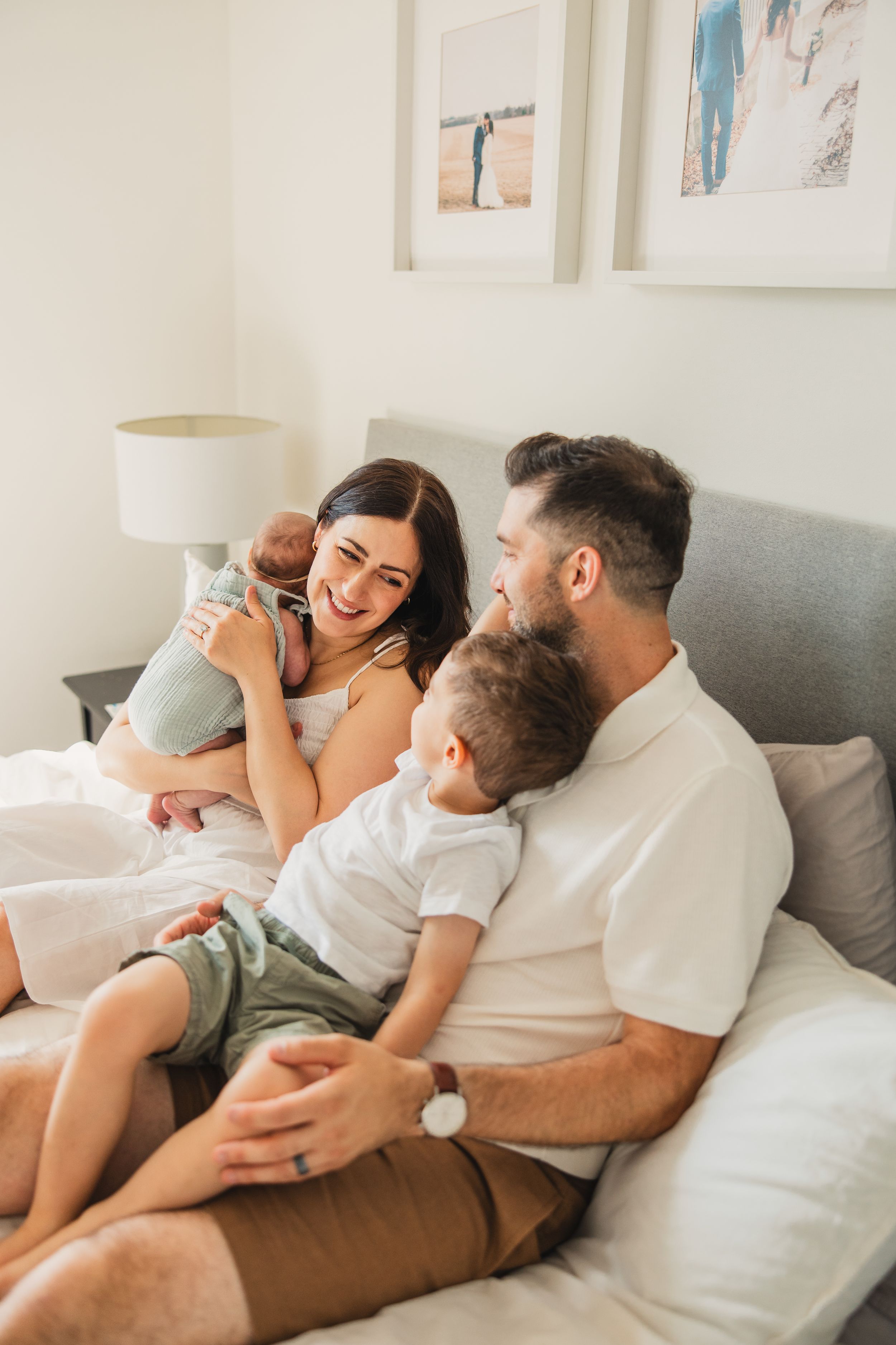 A family of four lounges together in bed with white bedding and pillows while sharing a relaxed and intimate moment.
