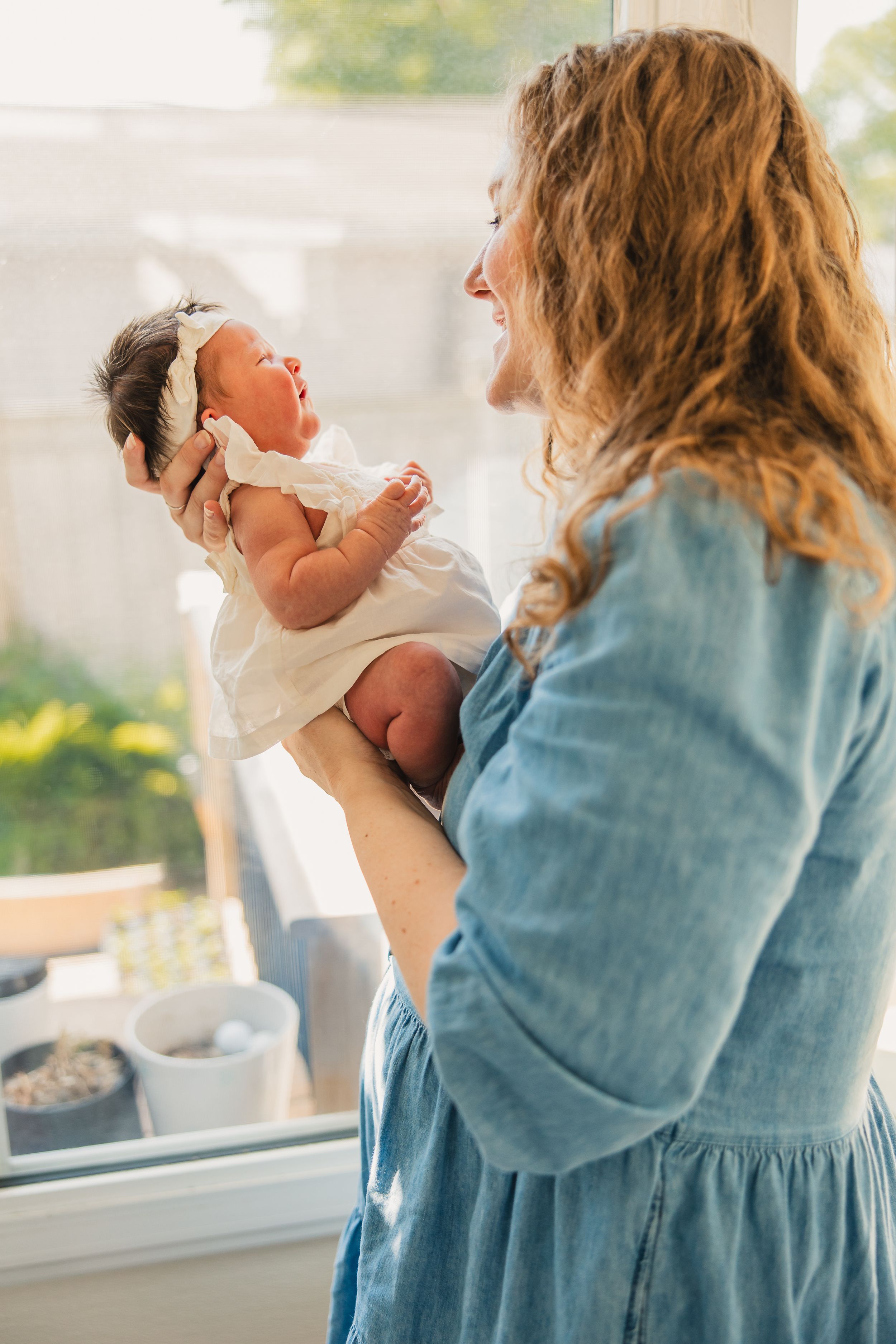 A woman in a blue denim dress holds a smiling baby wearing white, both sharing a joyful moment by a window.