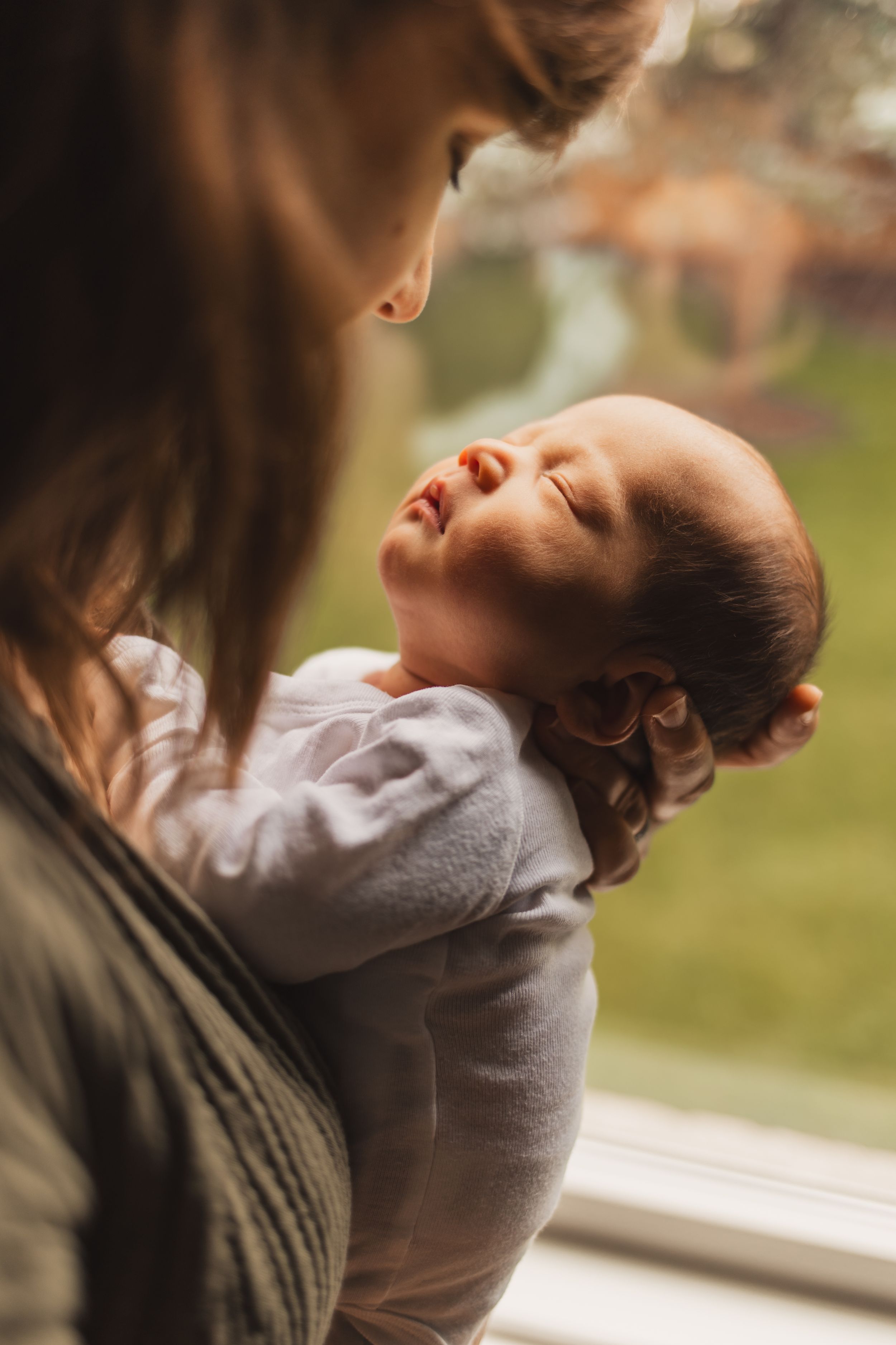 A tender moment captured as a parent cradles a sleeping baby in a soft white onesie near a window.