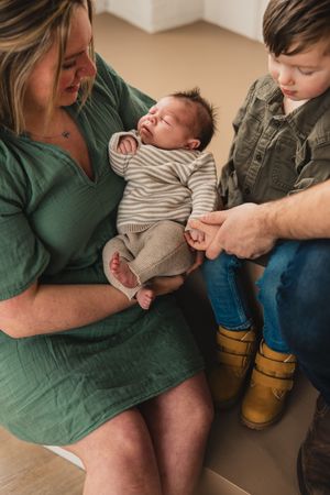 A newborn baby in striped onesie rests peacefully on his parent's lap while his brother wearing yellow boots sits nearby holding his hand.