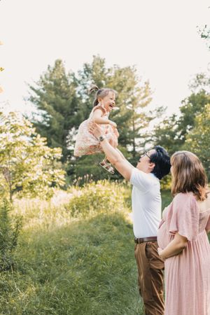 A family playfully jumps together in a sunlit meadow with pine trees in the background, wearing pink and brown attire for a maternity photo.
