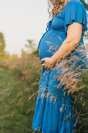 A pregnant woman in a flowing blue dress stands in a field of tall grass during golden hour.