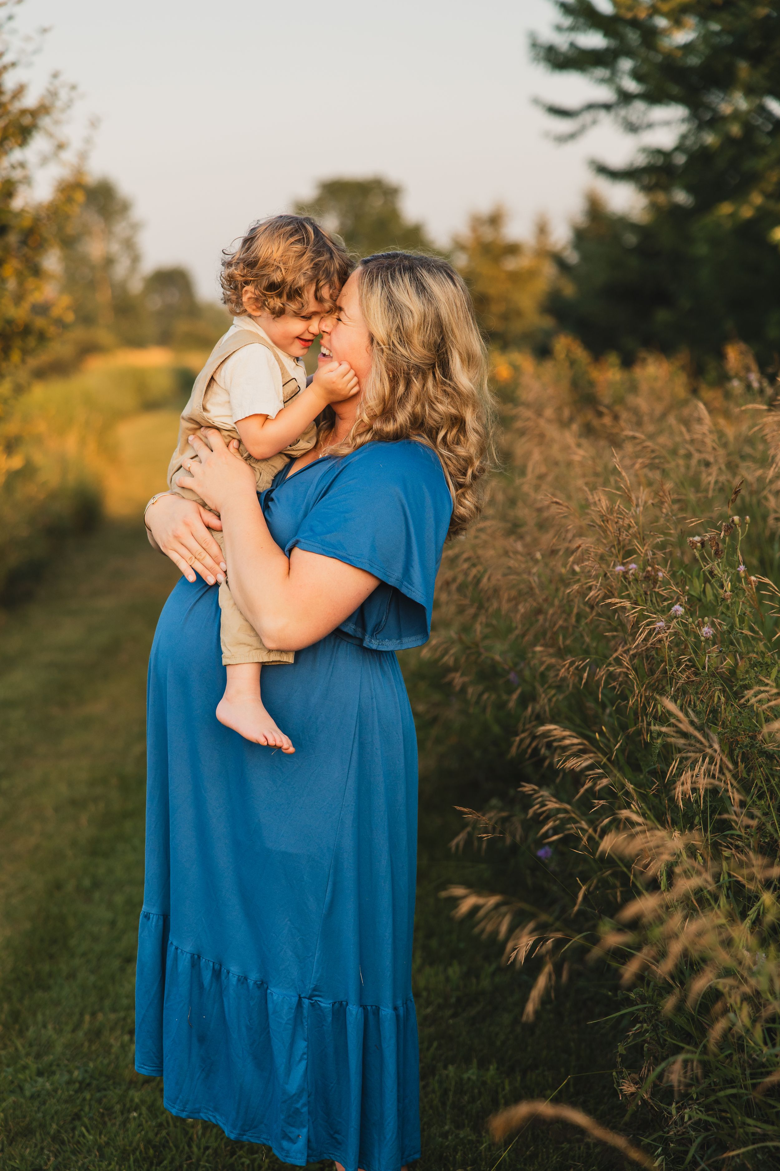 A pregnant person wearing a blue dress holds a small child in a golden-lit field during sunset.