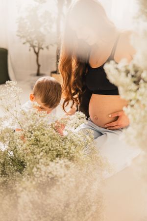Pregnant mother sits next to child in soft sunlight among delicate white baby's breath flowers creating an ethereal and dreamy atmosphere.