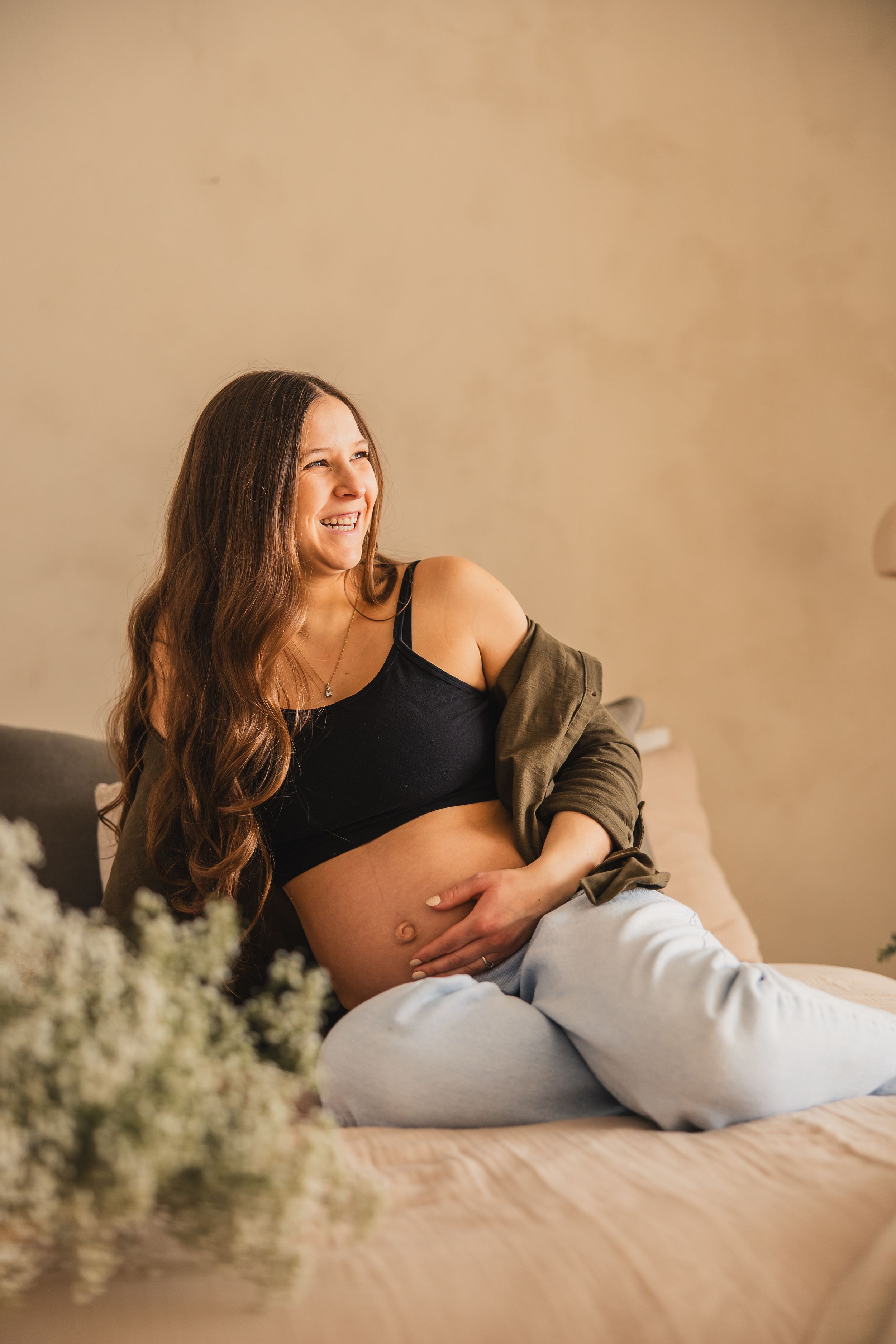 Maternity photo series showing someone in black top and white pants reclining on bed with soft beige lighting.
