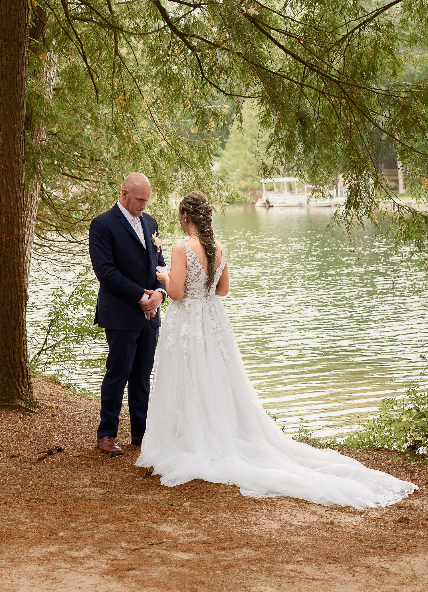 A bride and groom share a private moment by a serene lake under towering evergreen trees.