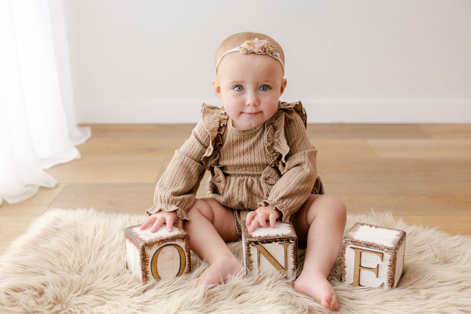 Small baby wearing a white tank top with "ONE" text sits on hardwood floor against white wall.