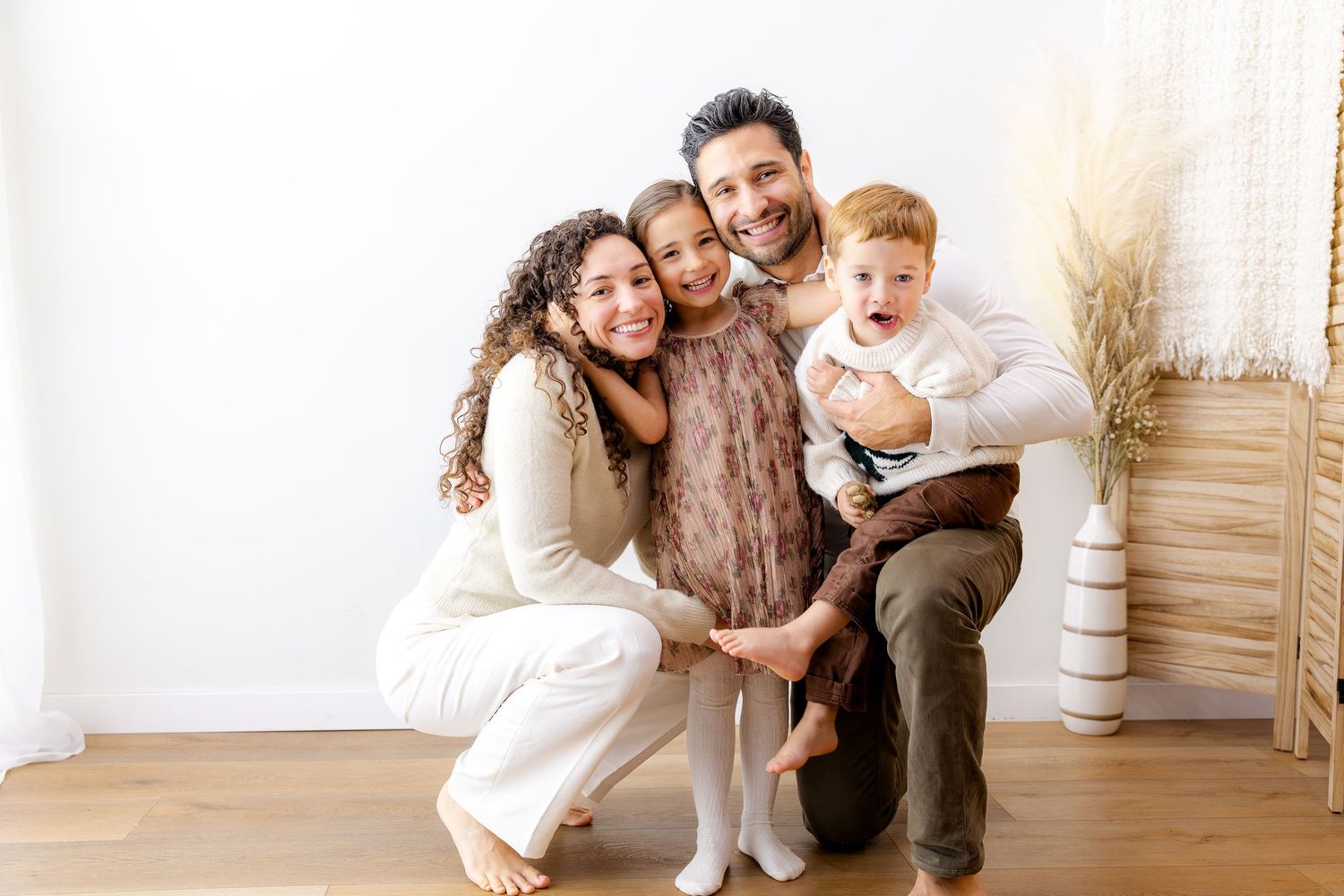 A family of four, with two young boys laughing joyfully, sits outdoors among tall dried grass at sunset.