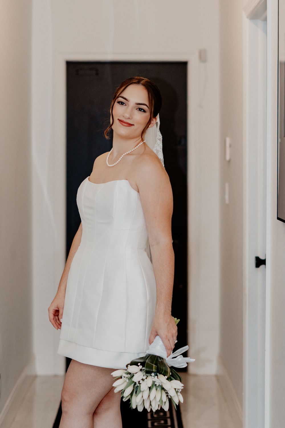 Bride in a white dress stands in a hallway holding a bouquet of white flowers.
