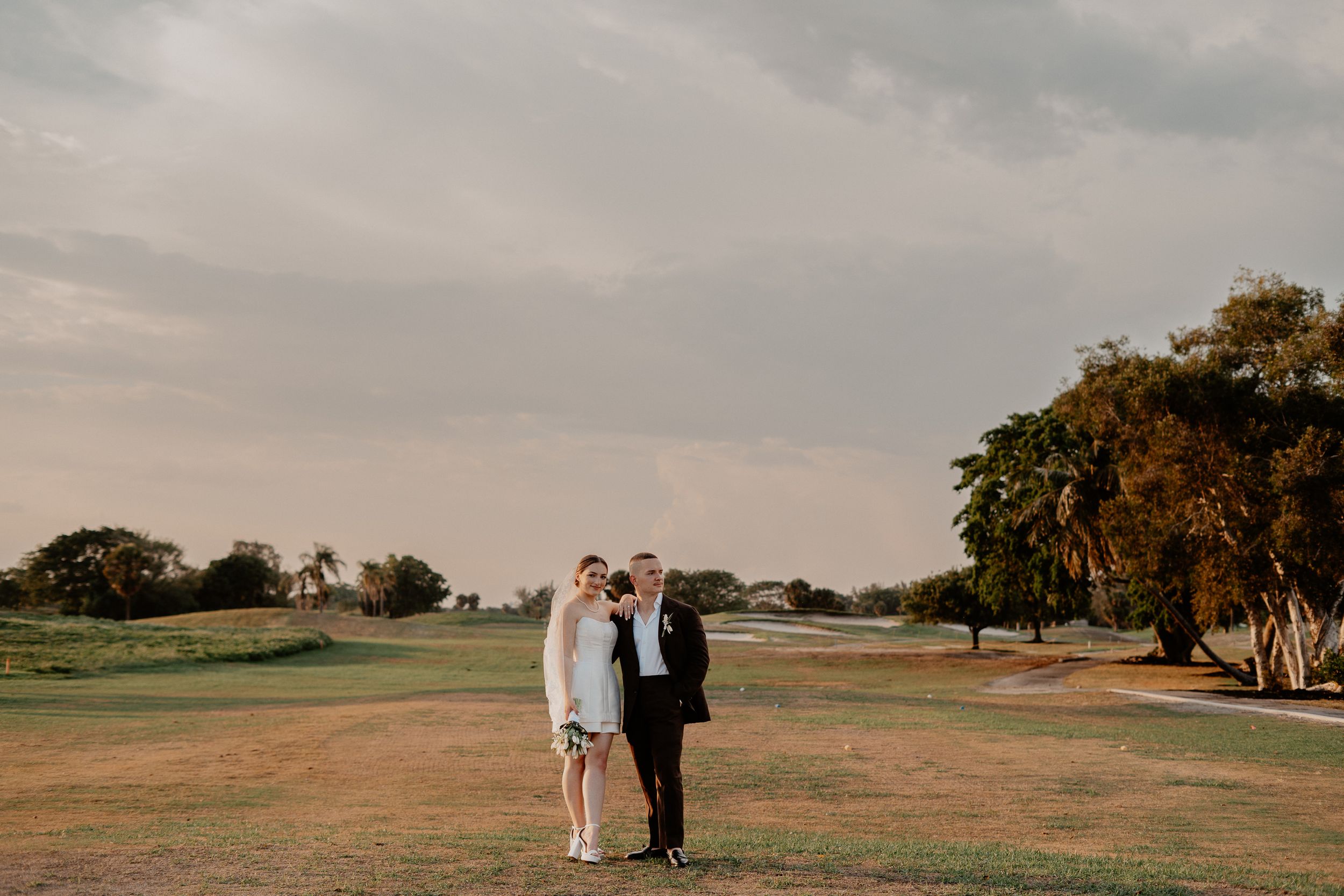 A couple stands together on a scenic golf course at sunset with oak trees in the background.
