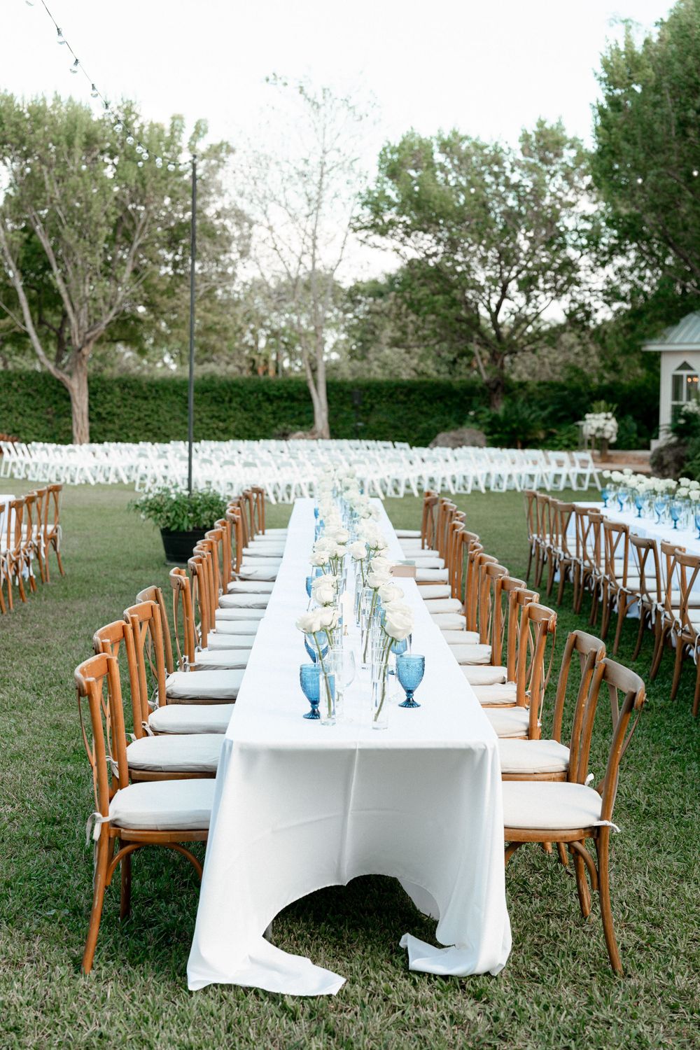 Long banquet table with wooden chairs and white linens set up for an elegant outdoor garden wedding reception.