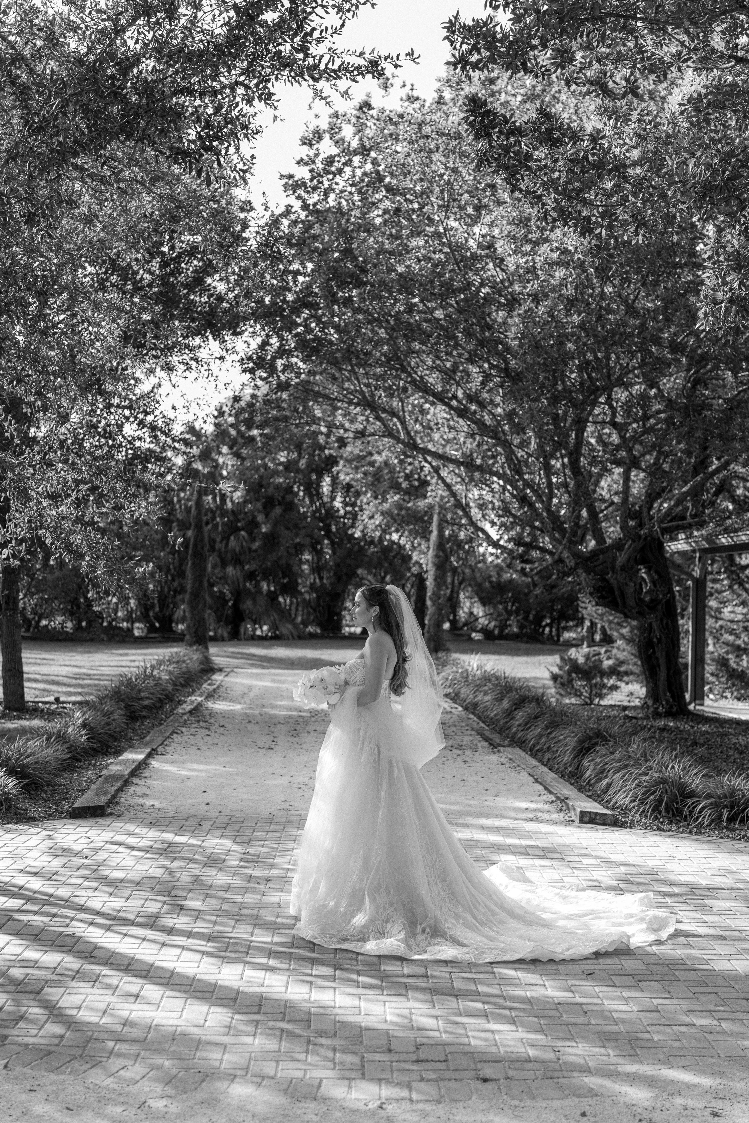 A bride in a flowing white wedding gown twirls gracefully on a brick pathway surrounded by lush trees and greenery.