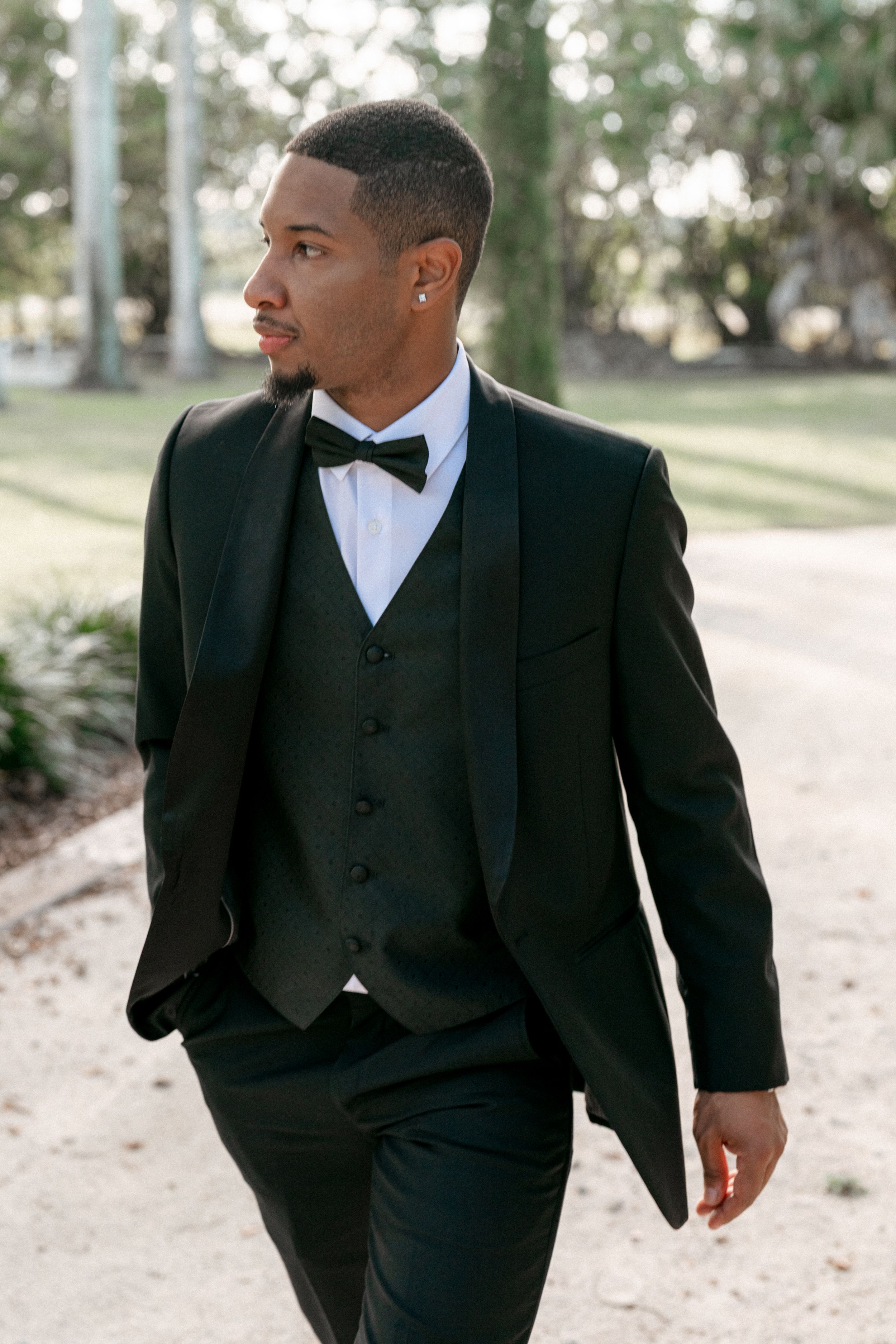 A groom in a classic black tuxedo and bowtie poses elegantly in an outdoor garden setting.