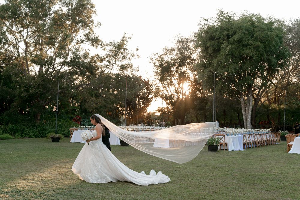 Bride's veil flowing dramatically in the wind at an outdoor garden wedding reception setup at sunset.
