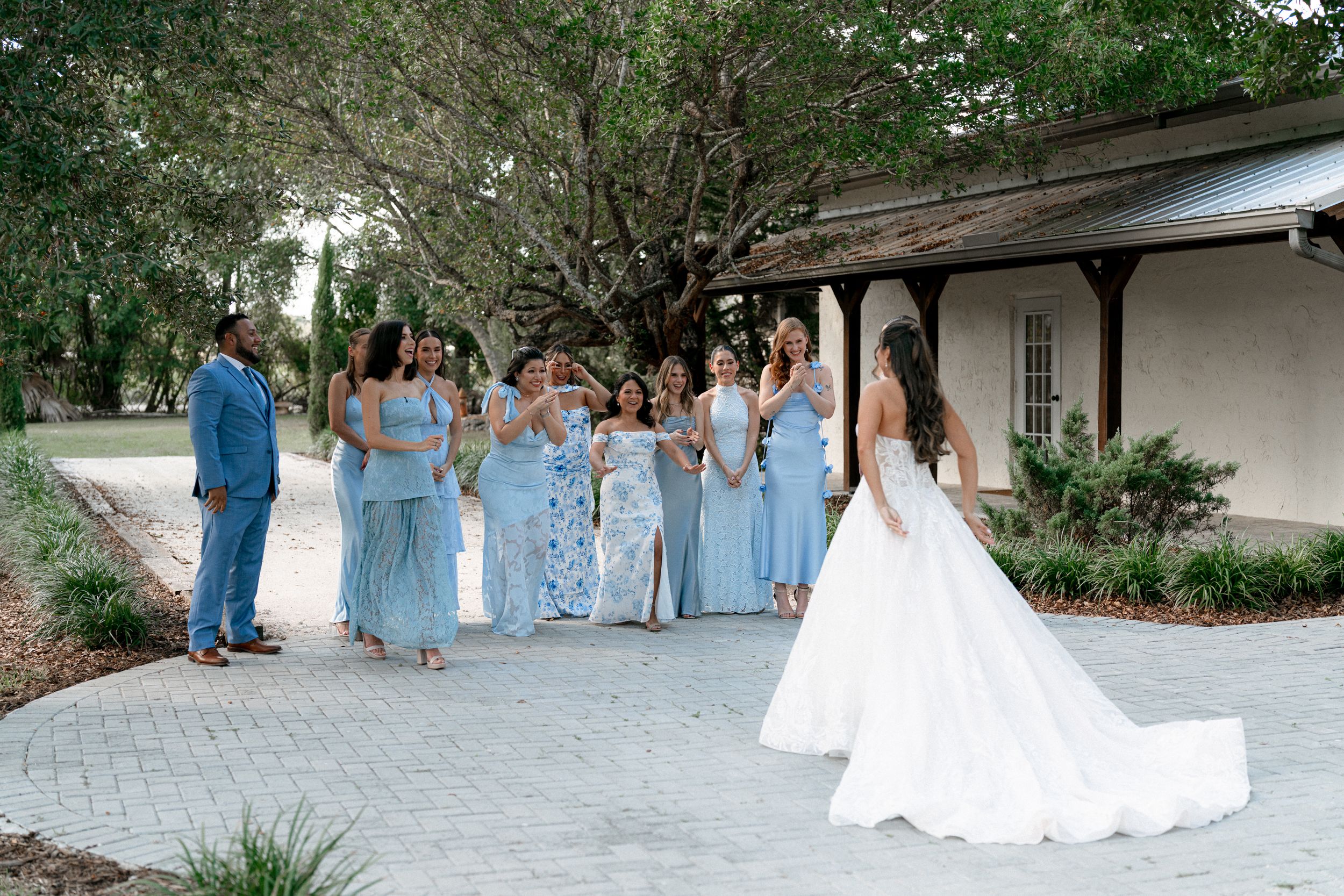 Wedding party in light blue dresses walking outdoors along a garden pathway during a sunny day.