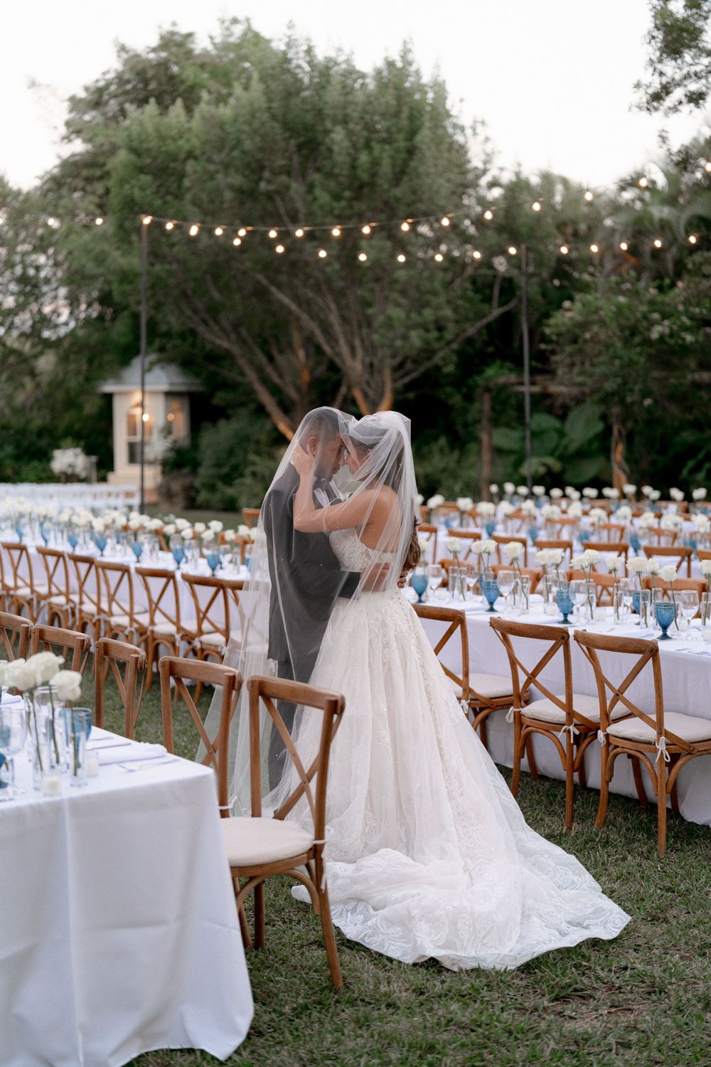 Outdoor wedding reception setup with wooden cross-back chairs, white tablecloths and string lights in a garden setting.