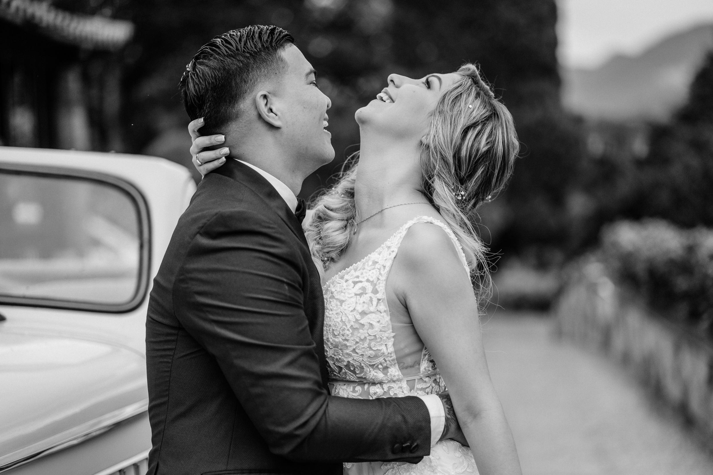 A romantic black and white wedding photo of a couple sharing a passionate moment beside a vintage car.