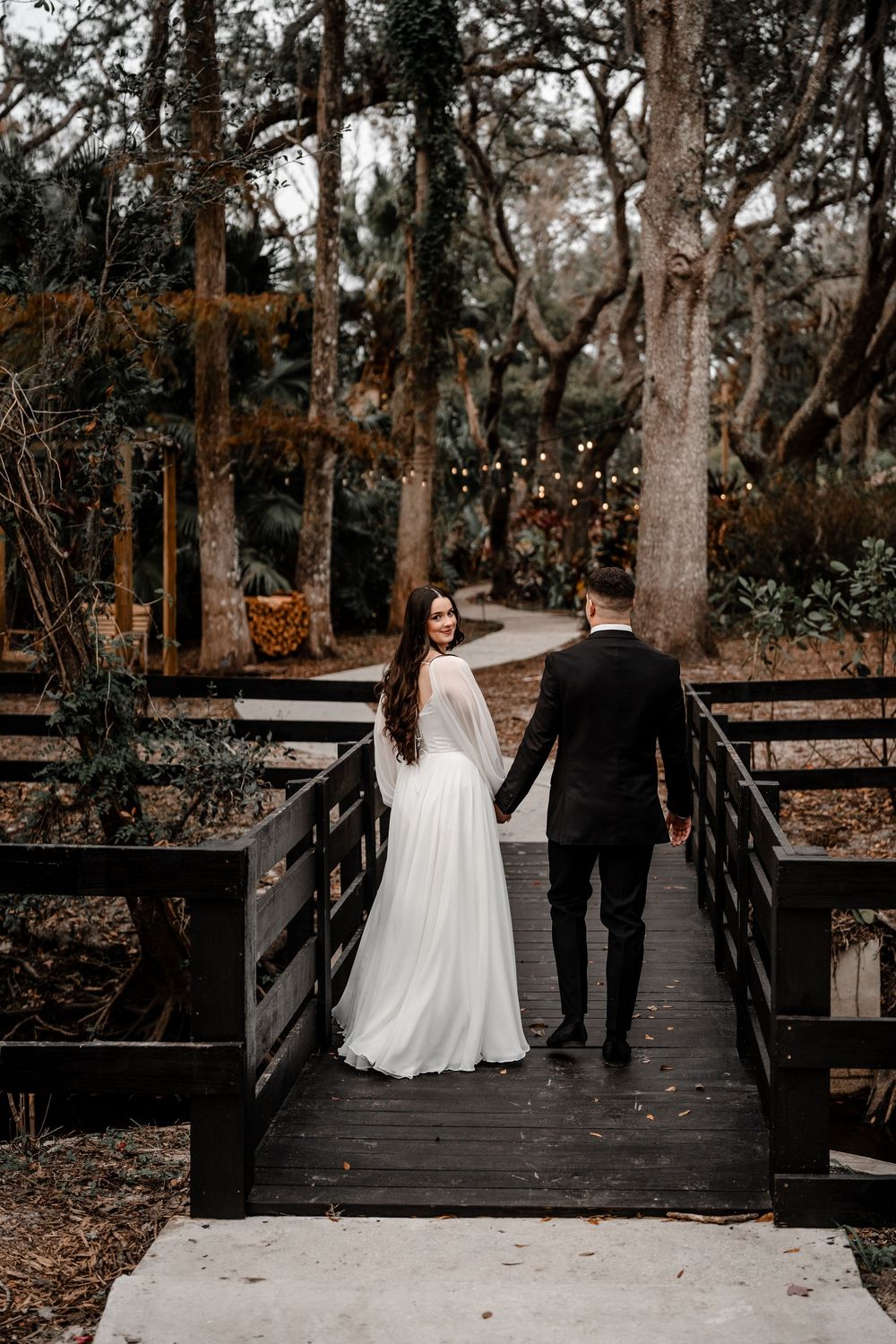 A couple in wedding attire walks together across a wooden bridge surrounded by trees in a natural outdoor setting.