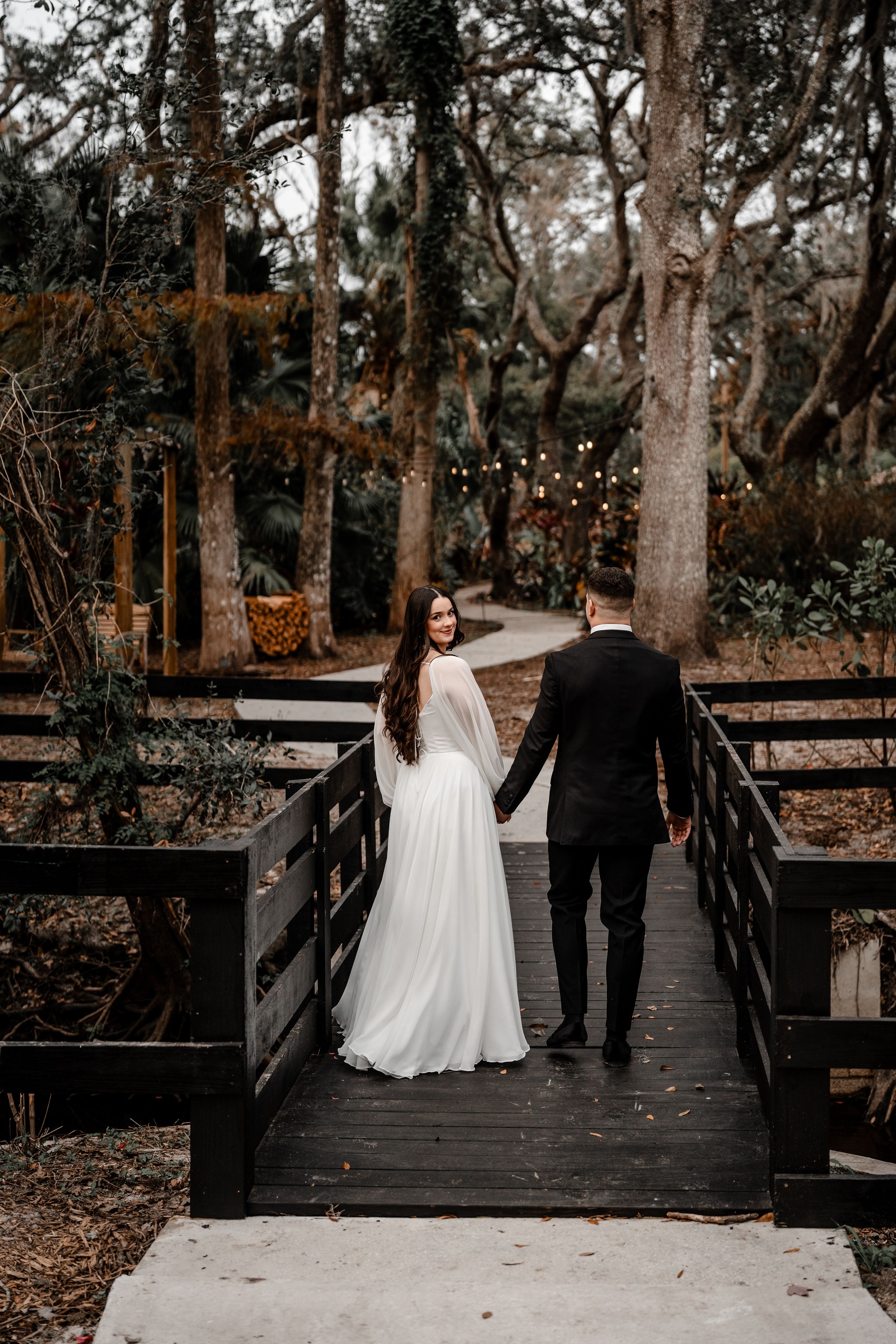 A couple in wedding attire walks together across a wooden bridge surrounded by trees in a natural outdoor setting.