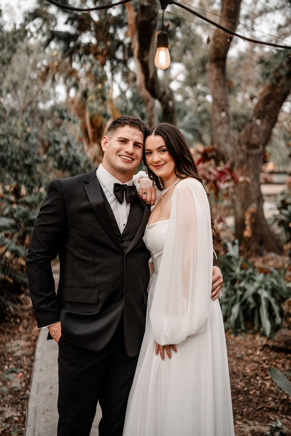A couple poses together outdoors under string lights at a romantic garden wedding ceremony.
