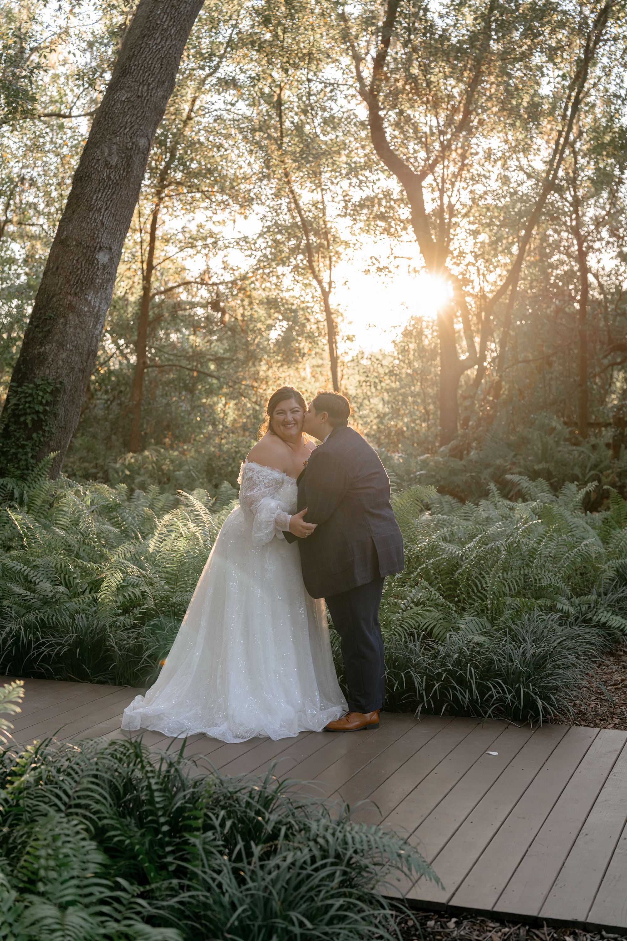 A couple shares an intimate moment on a wooden deck surrounded by ferns as golden sunlight filters through trees.