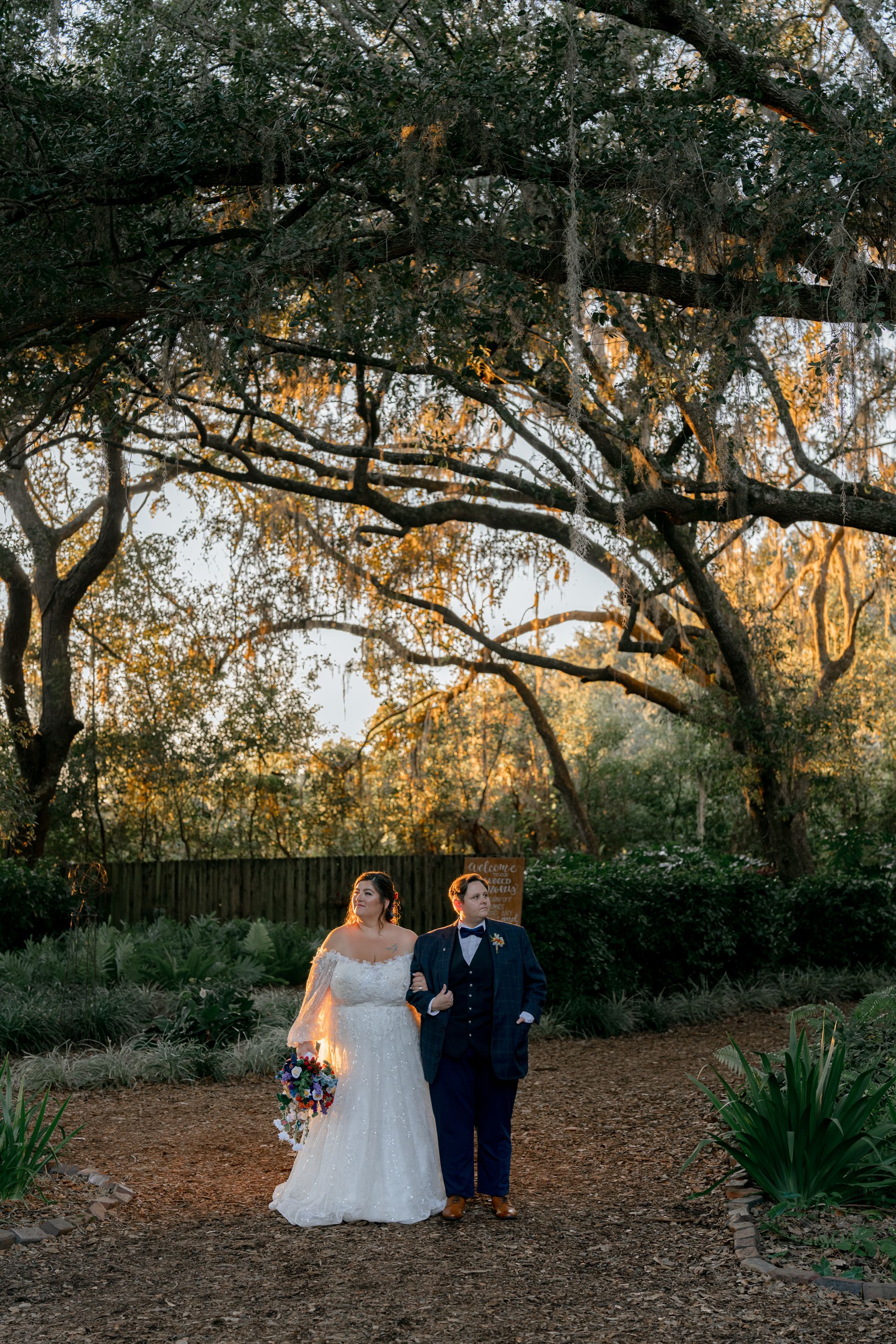 Couple in wedding attire standing under oak trees at sunset in a rustic outdoor setting.