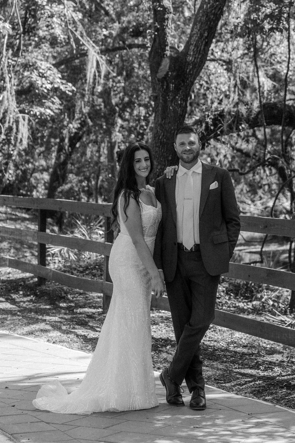 A couple in wedding attire poses together in a wooded outdoor setting with sunlight filtering through the trees.