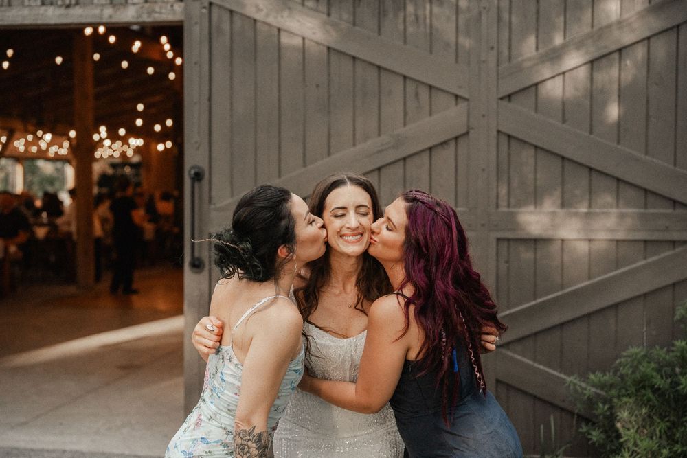 Three friends share a joyful embrace at a rustic venue decorated with twinkling lights.