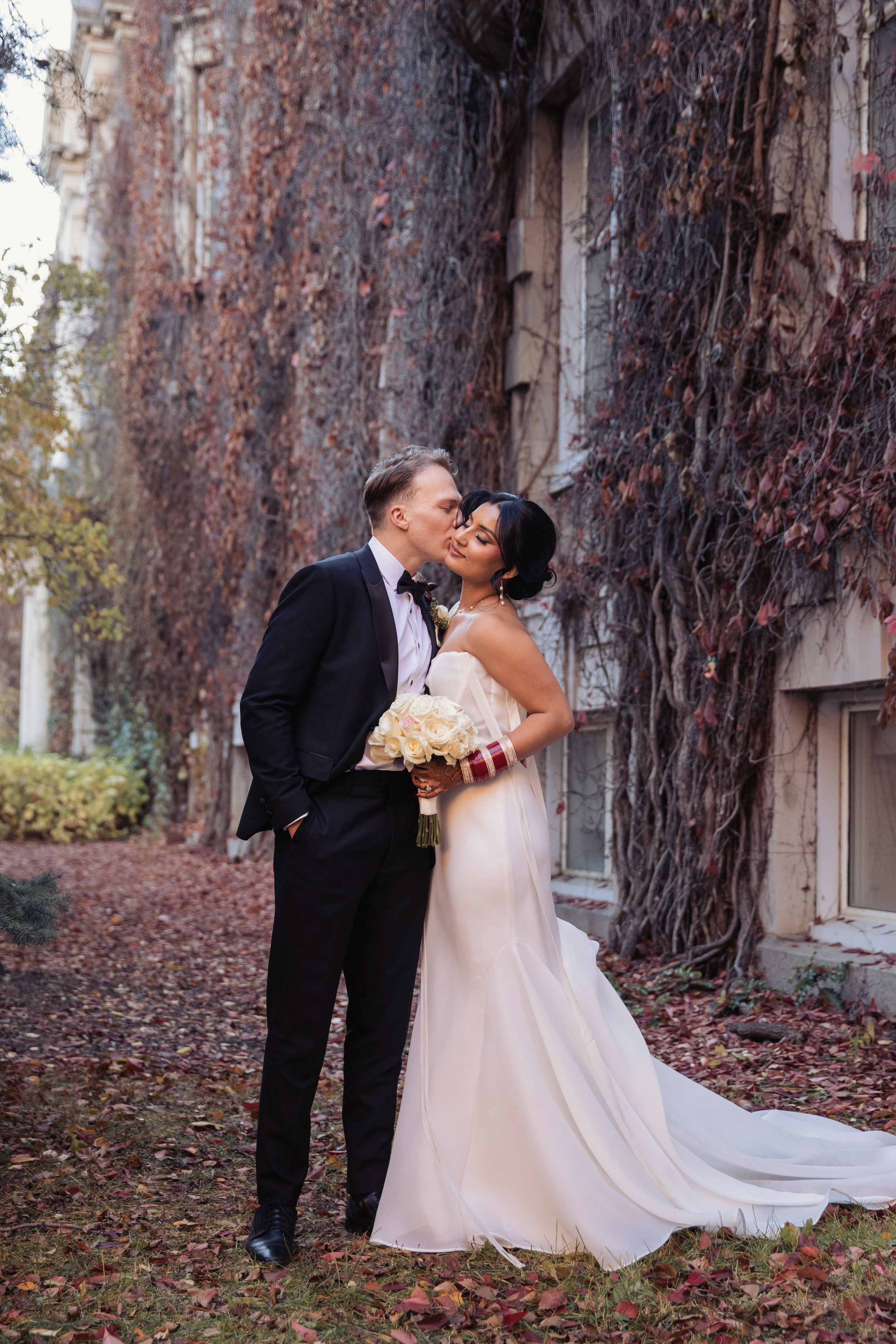 Bride and groom share a romantic kiss outdoors against an ivy-covered wall in autumn.