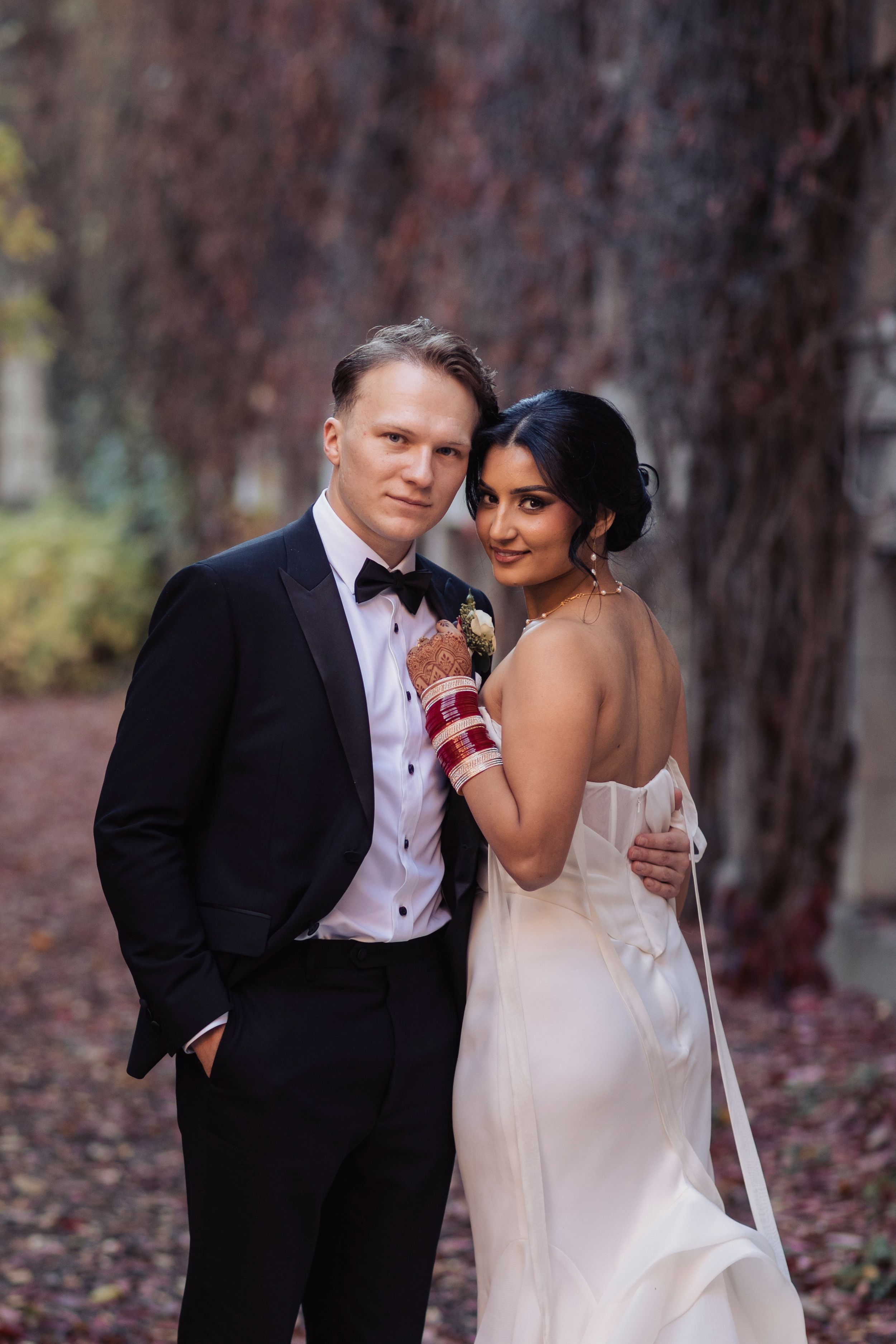 Elegant interracial wedding couple posing outdoors, groom in black tuxedo, bride in white backless gown with bridal bangles.