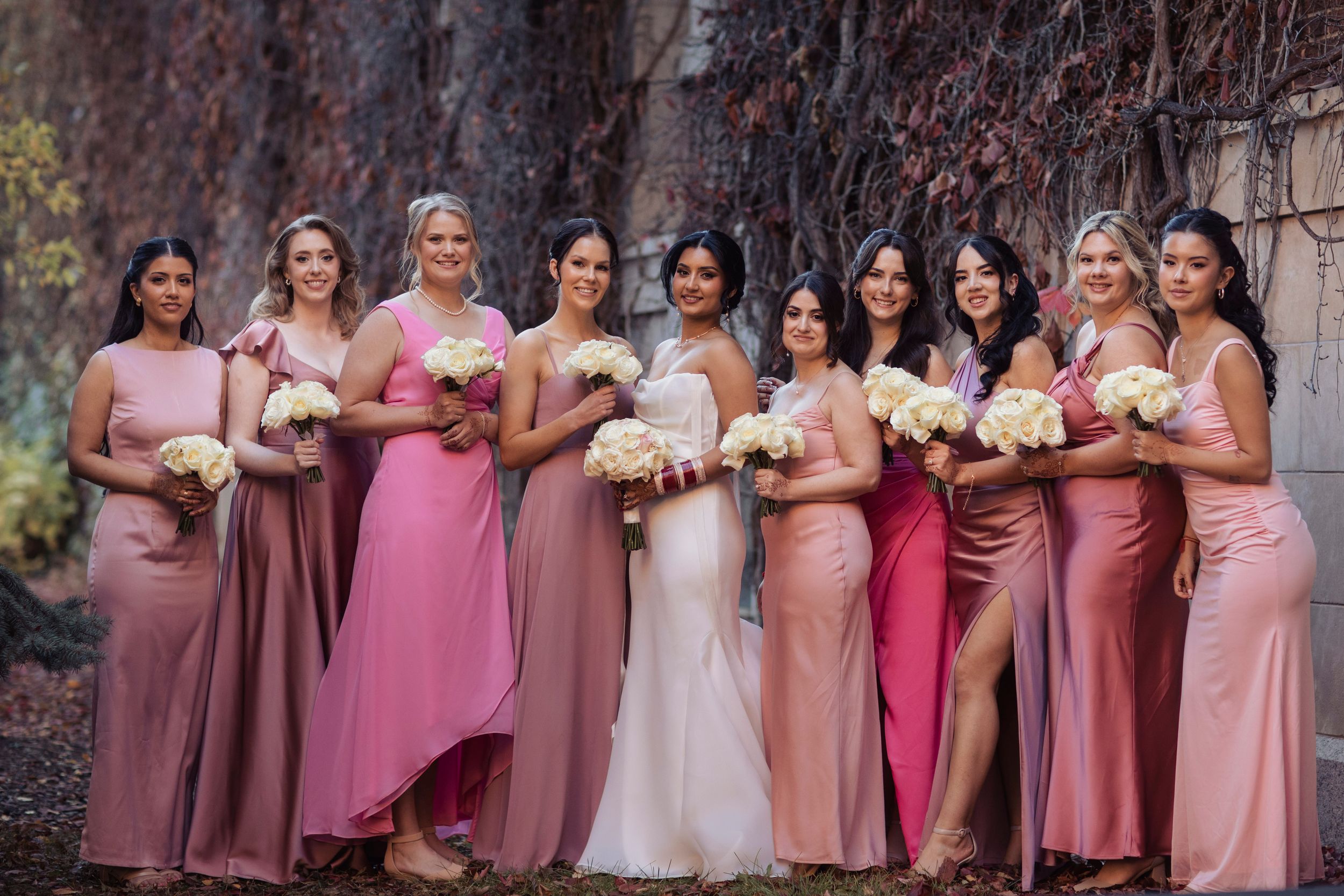 Bride in white gown poses with eight bridesmaids in pink dresses holding white bouquets outdoors.