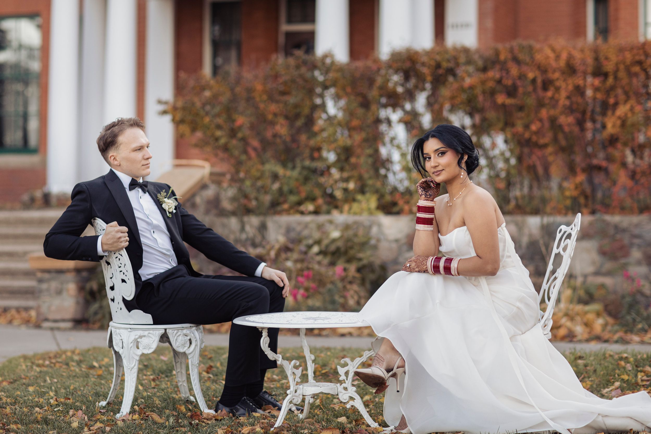 Bride and groom sitting at white iron table outdoors in fall, wearing wedding attire, elegant estate in background.