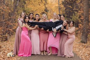 A group of people wearing pink and mauve dresses pose together in a scenic autumn forest setting.
