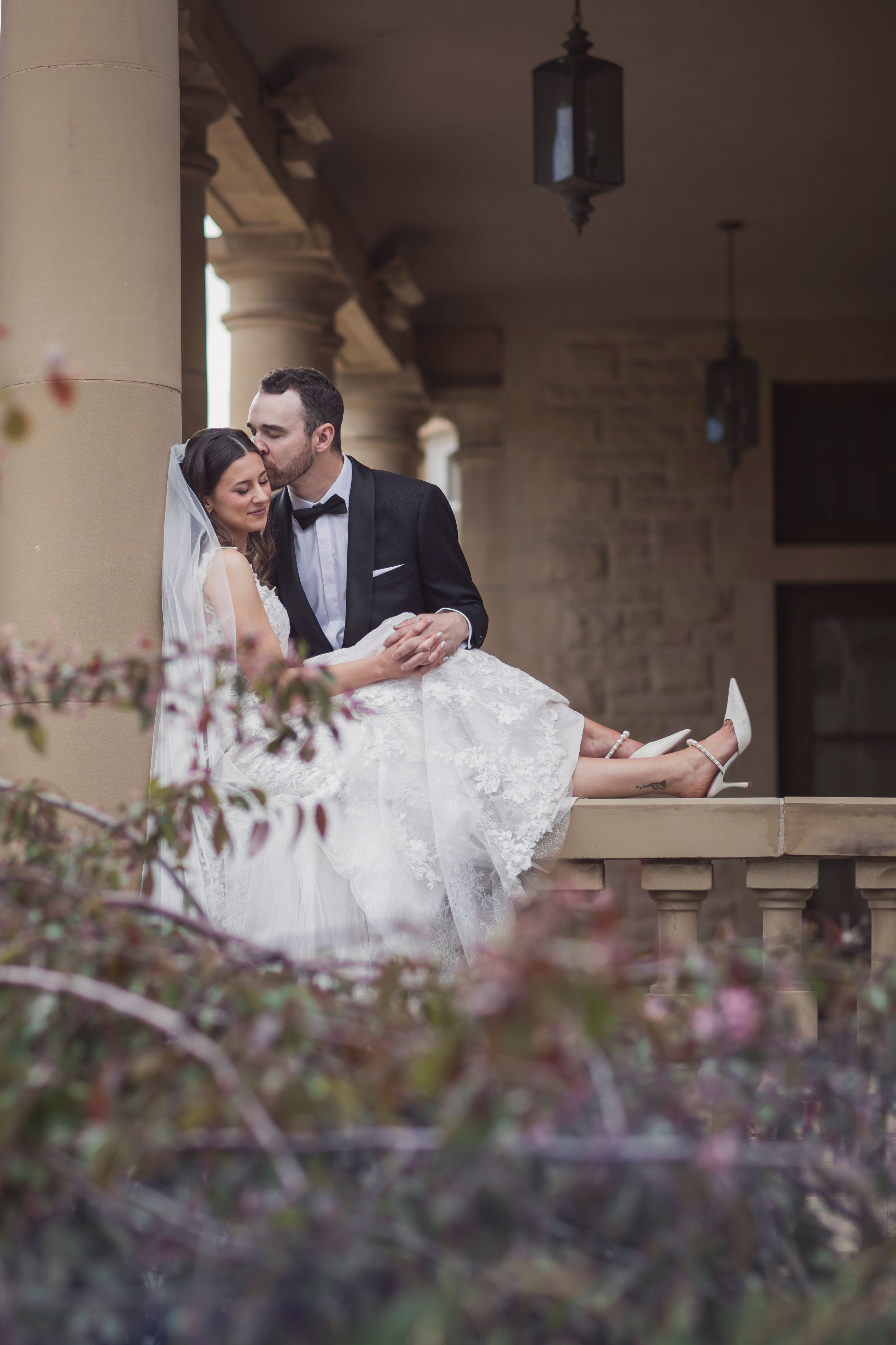 Bride and groom sit intimately on a stone balcony, sharing a tender moment in classic wedding attire.