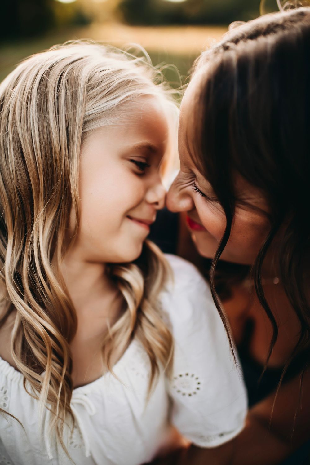 A mother and young blonde girl touch foreheads tenderly, sharing a sweet moment outdoors in warm golden light.