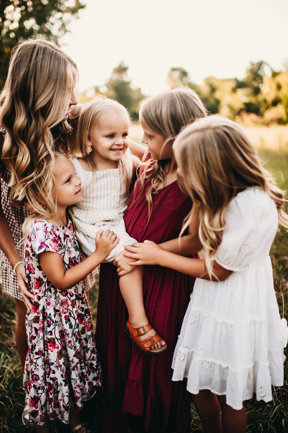 A mother and three young daughters share a joyful moment outdoors in golden light, wearing floral and neutral dresses.