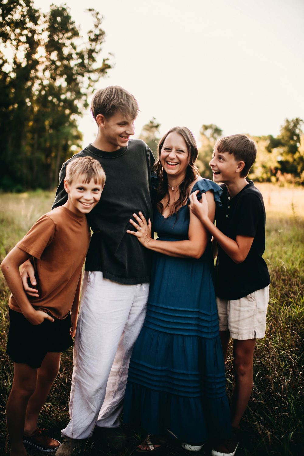 A smiling mother in a blue dress laughs with her three sons outdoors in a sunny field at golden hour.