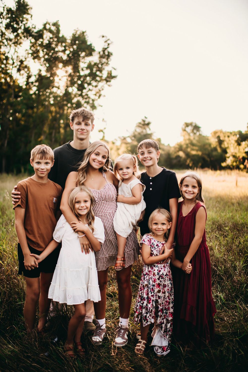 A woman poses with seven children of various ages in a sunlit field surrounded by lush green trees.