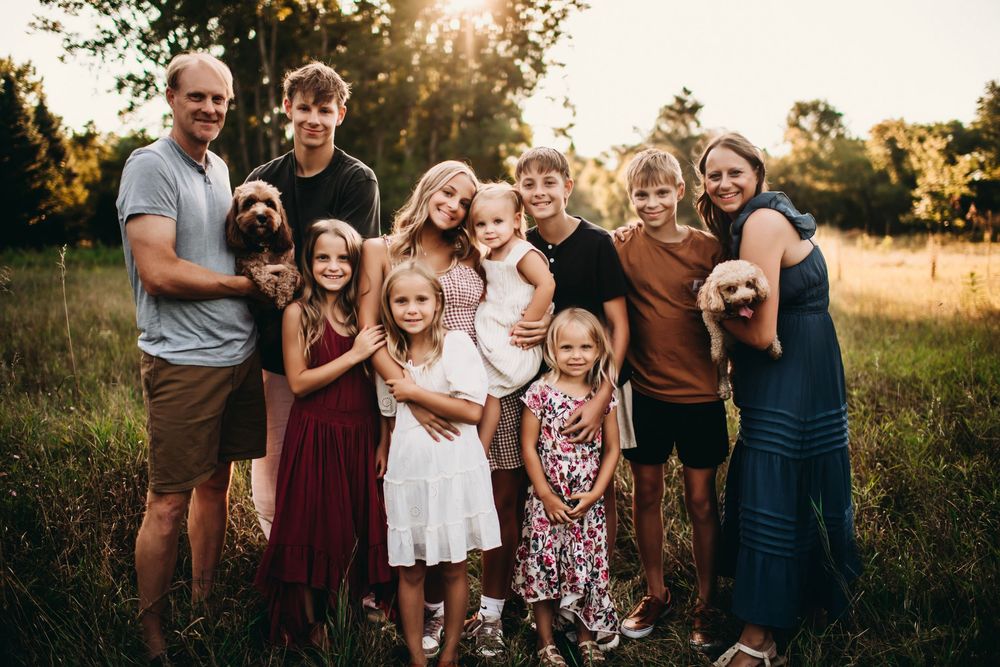 Large family with two dogs posing outdoors in a sunny field during golden hour for a professional portrait.