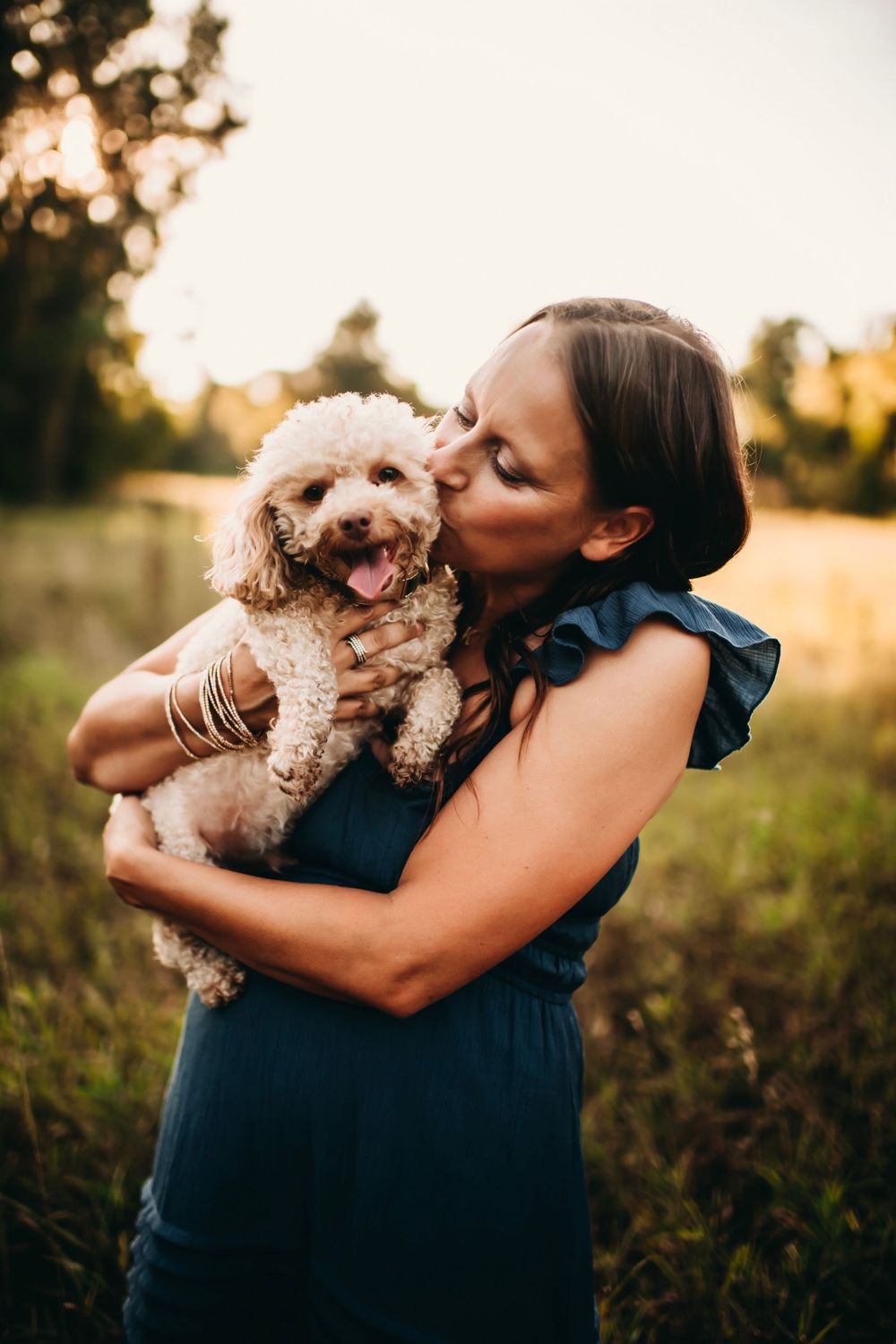 Woman in teal dress kissing her fluffy cream poodle in a sunlit field at golden hour.