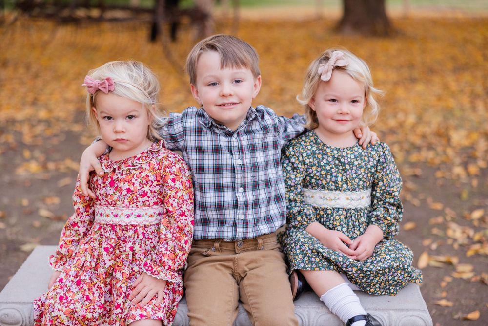 Close-up portrait of three children in patterned dresses and plaid shirt on wooden bench.