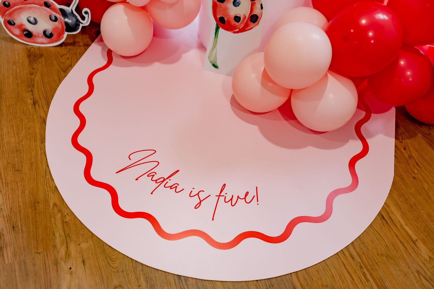 White circular placemat with red trim and cursive text displayed with pink and red balloon decorations.