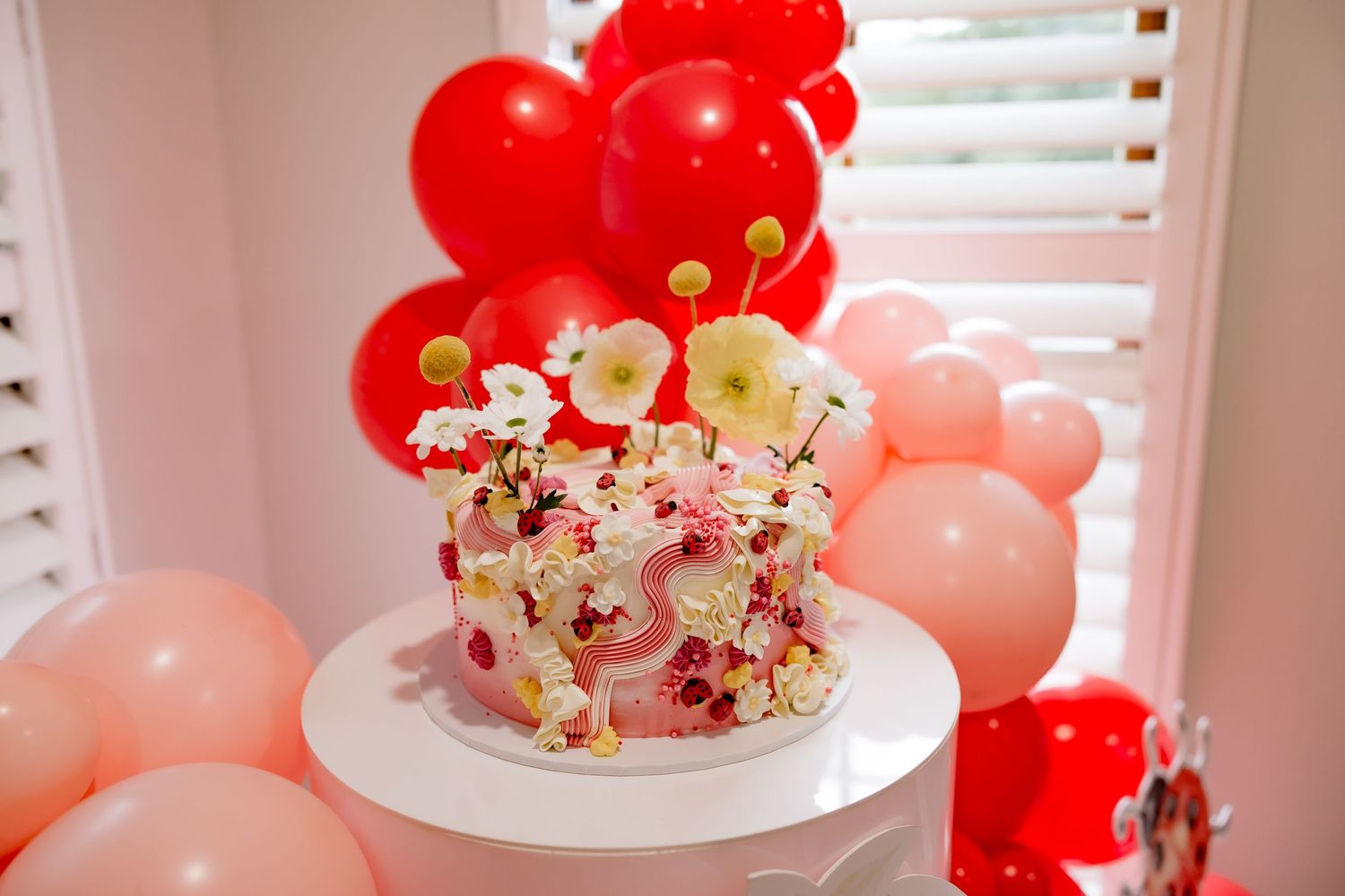 Elegant white cake decorated with pink flowers and red accents surrounded by red and pink balloons.