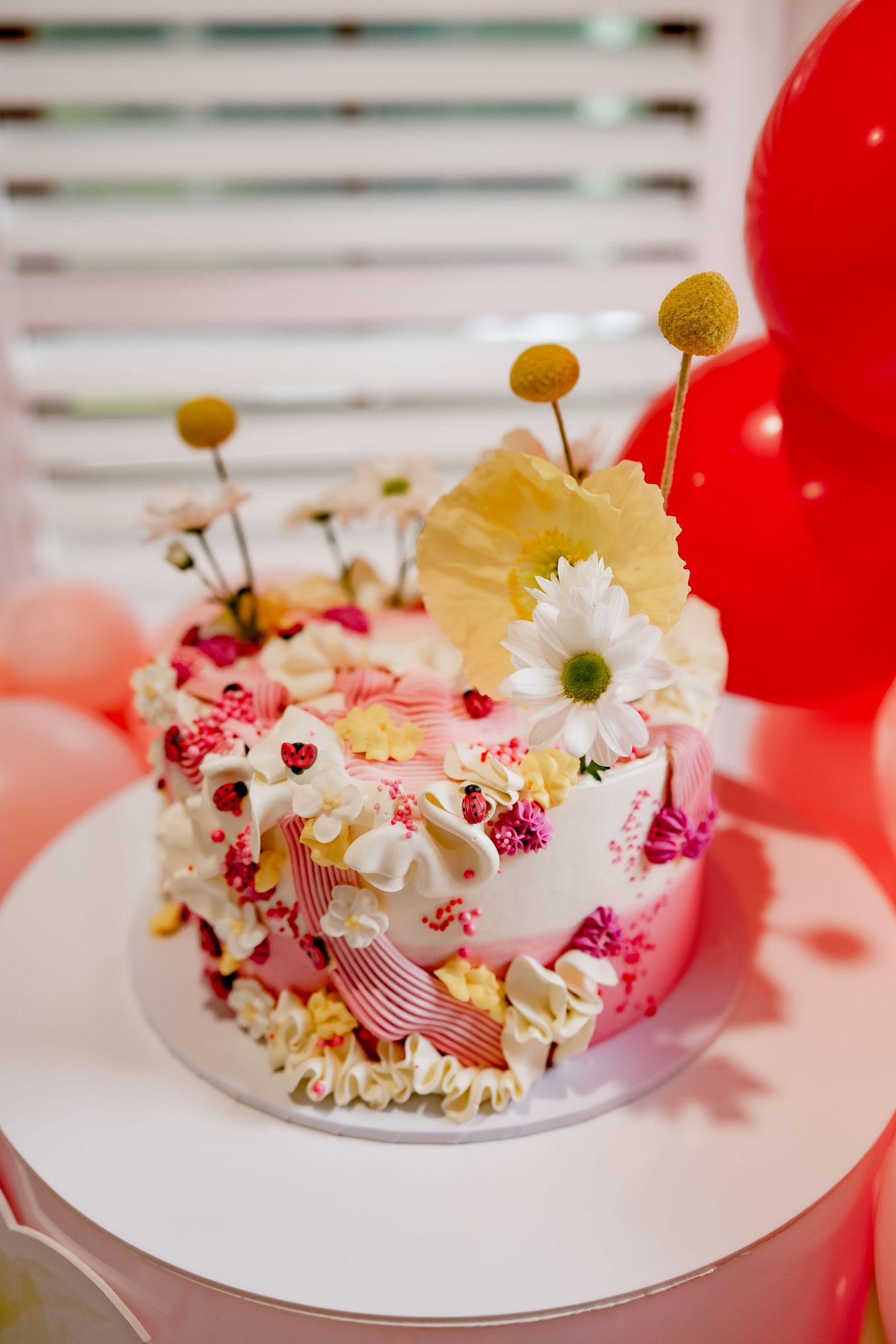 Whimsical cake topped with yellow craspedia balls and white daisies against pink and red balloon backdrop.