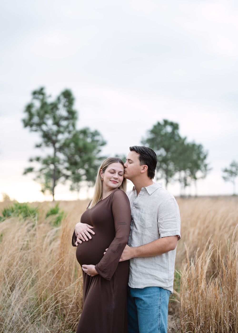 Pregnant couple embraces in a golden field at dusk, man kissing woman's cheek as she holds her bump.