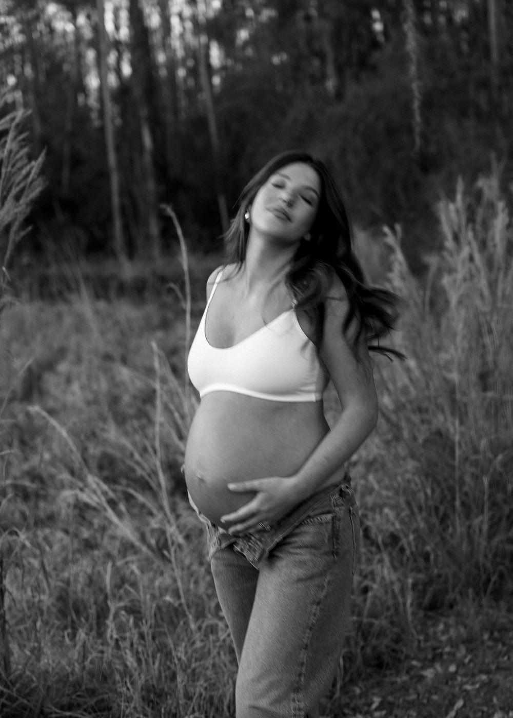Black and white maternity photo of woman in white bralette holding baby bump in tall field grass.