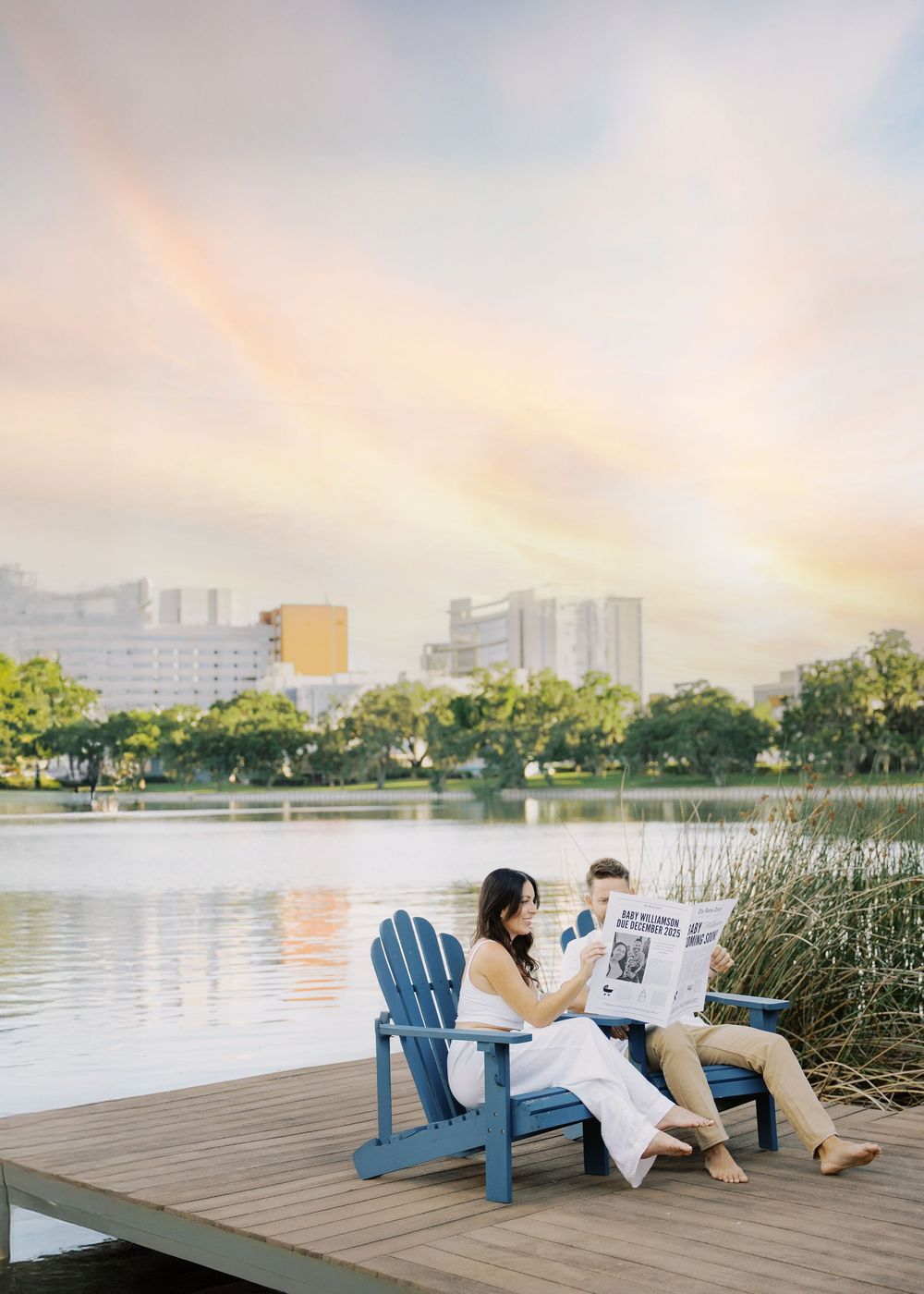 Couple sits in blue Adirondack chairs on lakeside dock reading newspaper under a rainbow sky.