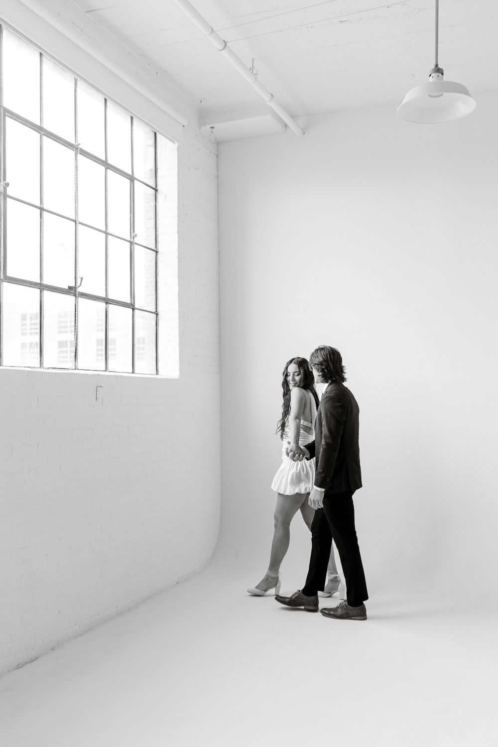 A minimalist black and white photograph of a couple standing in an empty white studio with large industrial windows.