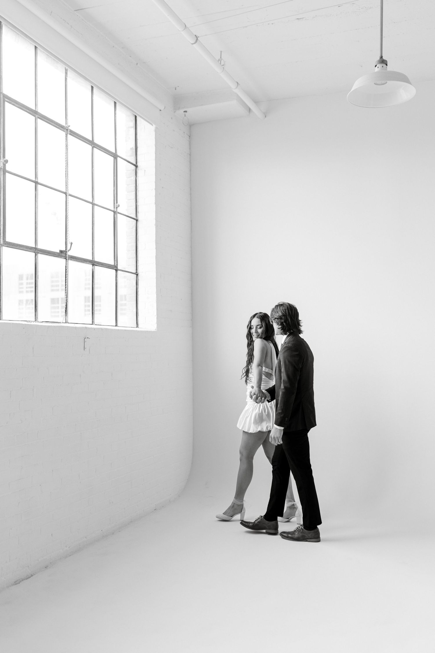 A minimalist black and white photograph of a couple standing in an empty white studio with large industrial windows.